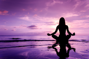 A woman meditating on the beach, by the shore surrounded by purple skies